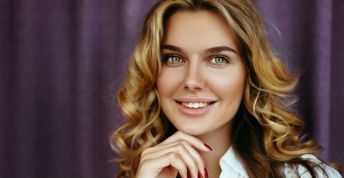 Portrait of a beautiful woman with curly blonde hair and hands under her chin, against purple drapes